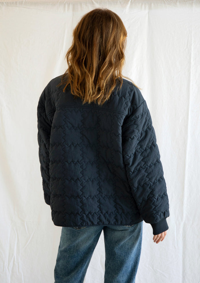 A woman wearing a navy blue jacket with squiggle shaped quilting and gingham and floral patchwork detailing at front.
