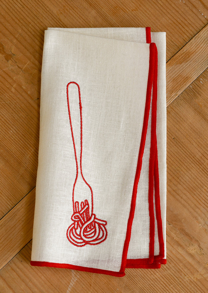 An ivory cloth dinner napkin with red edging, and red embroidery of spaghetti on fork.