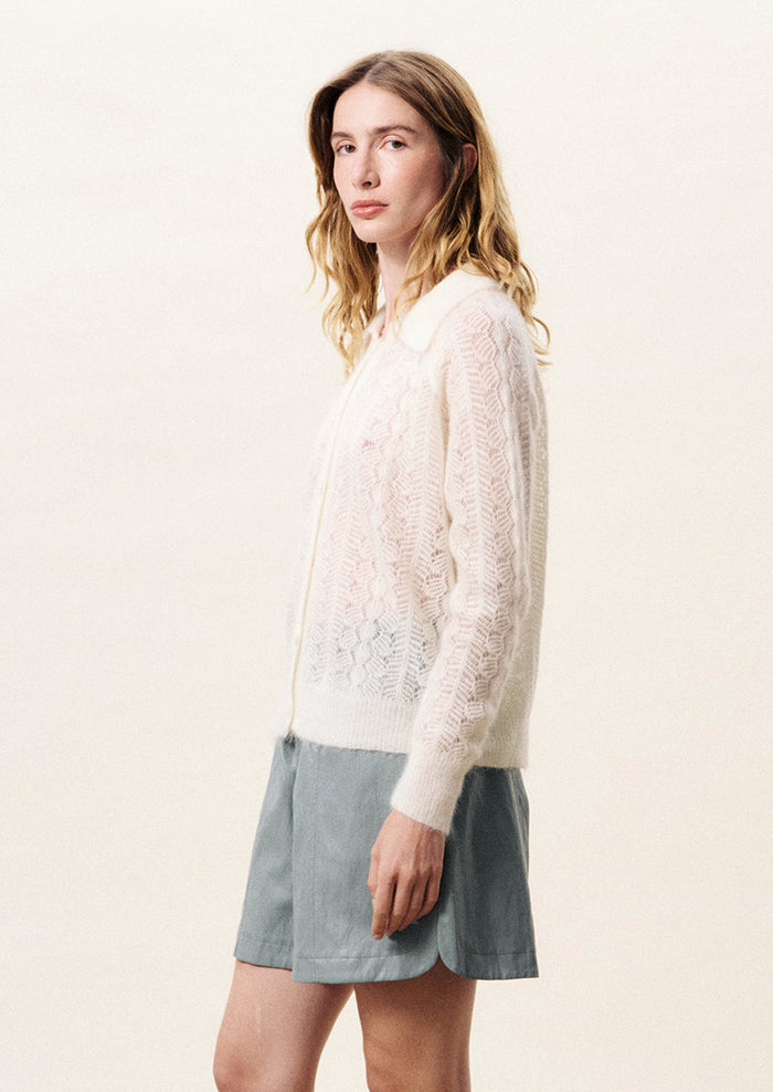 Person wearing a white lace cardigan with arms crossed on a light background