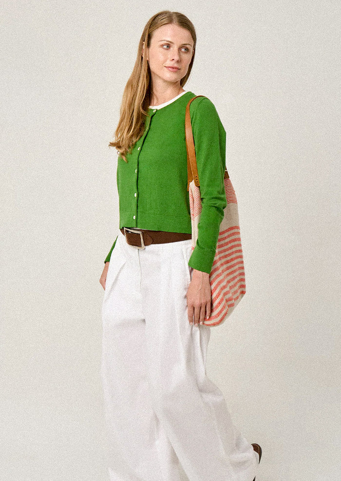 Woman wearing a green cardigan and white pants, holding a striped bag against a plain background