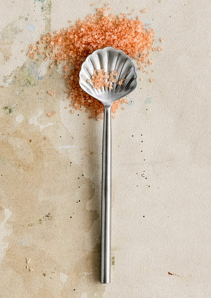 Metallic shell-shaped spoon with pink sea salt on a textured beige surface